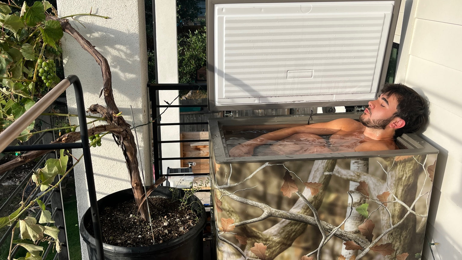 Person lying inside an open CryoConverted plunge with camouflage design, surrounded by plants and outdoor elements.